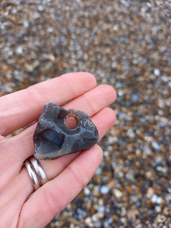A hand holding a hag stone against a pebble beach backdrop